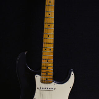 Black Fender Stratocaster with white pickguard and maple neck, shown from headstock to body with its vibrato arm against a dark background.
