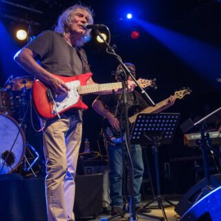 Albert Lee singing while playing a red electric guitar, with his band behind him.