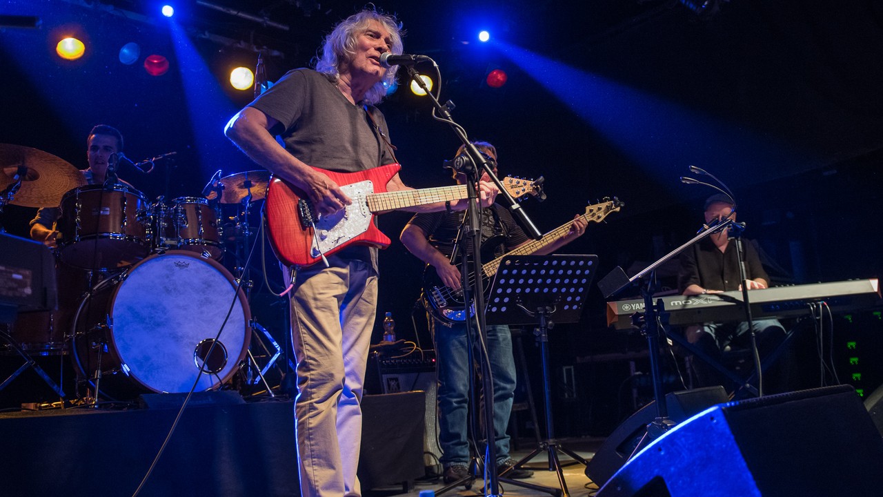 Albert Lee singing while playing a red electric guitar, with his band behind him.