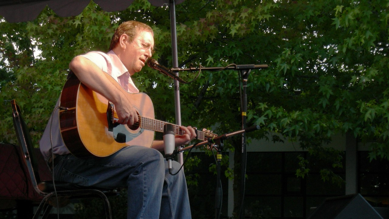 Bert Jansch seated outdoors, singing while playing an acoustic guitar at Bumbershoot.