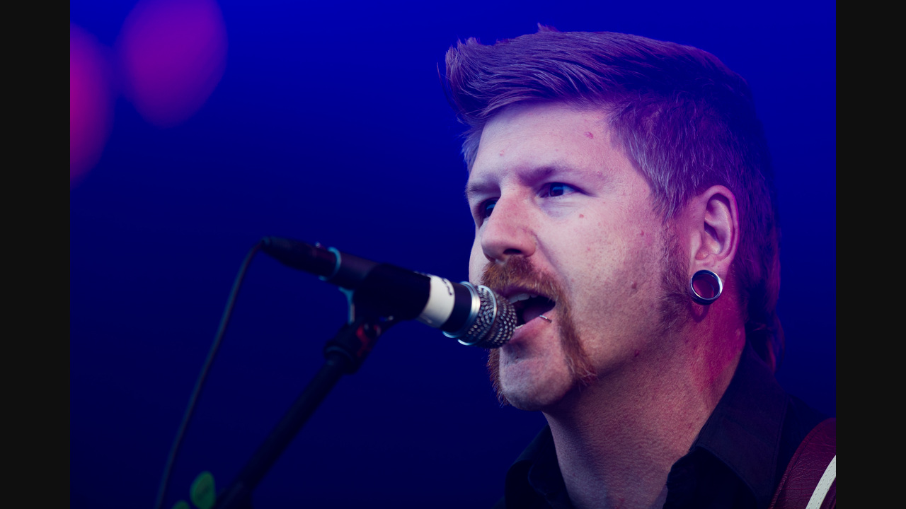 Bill Kelliher in a close stage portrait at a microphone under blue and purple lights. His complete head is visible; no guitar appears in this portrait.