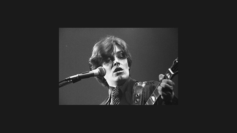 Black-and-white performance portrait of Bill Nelson beside a microphone, with part of a guitar neck visible.