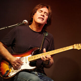 Carl Verheyen playing an electric guitar on stage, with both hands and the guitar visible.