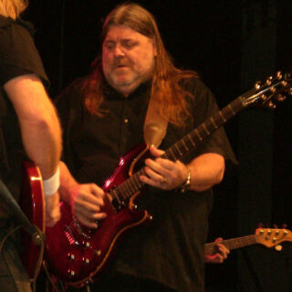 Dave Hlubek playing guitar on stage under warm concert lighting, with another performer partly visible in the foreground.