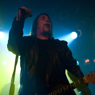 Dave Wyndorf holding a microphone on stage with a guitar partly visible under blue and green lights.