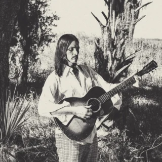 Duane Betts playing an acoustic guitar outdoors in a black-and-white promotional photograph.