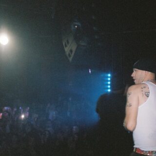 Archival photograph of fish narc in left-facing profile onstage, wearing a black head covering and white sleeveless shirt.