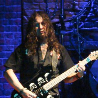 Michael Wilton playing a skull-graphic double-neck guitar on stage.