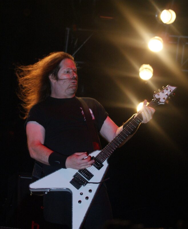 Gary Holt with long hair swept sideways playing a white Flying V-shaped guitar on stage