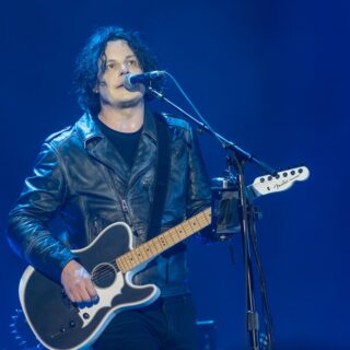 Jack White in a black leather jacket singing and playing a dark guitar under blue stage lighting at the Troxy in London.