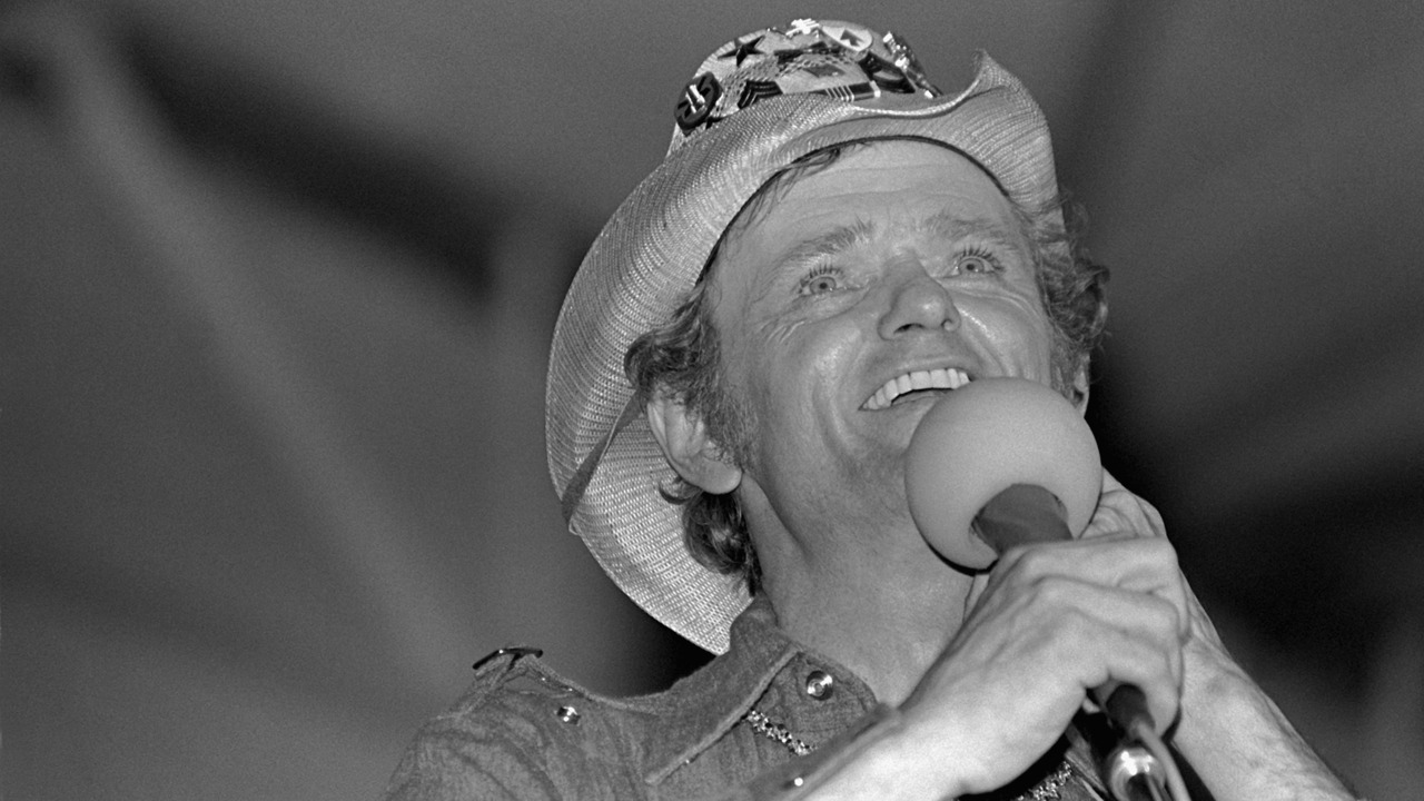 Jerry Reed smiling into a microphone while wearing a decorated hat.