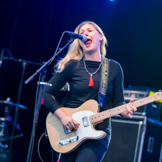 Joanne Shaw Taylor performing in the complete published concert photograph, with full head, both hands and entire principal guitar body, neck and headstock visible, within dark side margins.