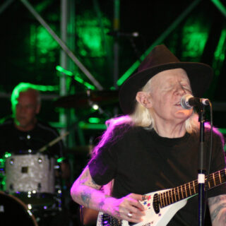Johnny Winter in a black hat, singing into a microphone while playing guitar, with a drummer behind him.