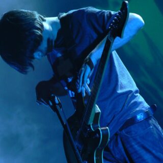 Jonny Greenwood leaning over a dark electric guitar under blue stage lighting.