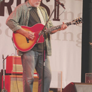 Jorma Kaukonen performing with Hot Tuna in 2009, standing with a red-and-sunburst guitar and both hands visible.