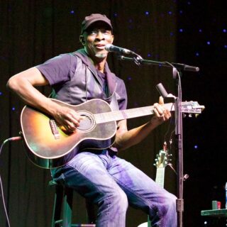 Keb’ Mo’ seated onstage, singing and playing a sunburst acoustic guitar.