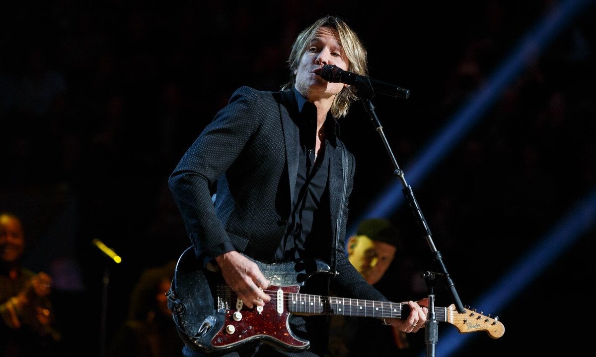 Keith Urban sings at a microphone while playing a fully visible worn dark electric guitar, with his complete head and both hands inside the frame. Blue stage lights cross the dark background.