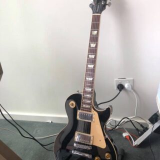 Complete black-finish Gibson Les Paul Standard on a stand, with its headstock, neck, cream pickguard and two humbuckers visible.
