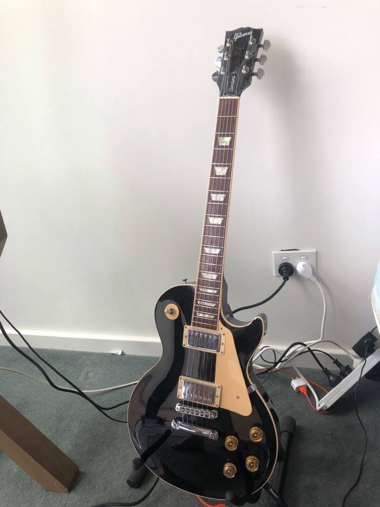Complete black-finish Gibson Les Paul Standard on a stand, with its headstock, neck, cream pickguard and two humbuckers visible.