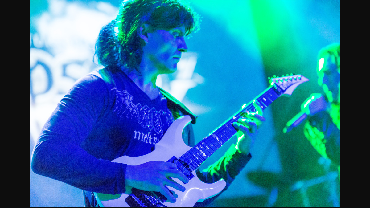 Luca Turilli playing a white electric guitar under blue and green stage lighting, with both playing hands and the headstock visible.