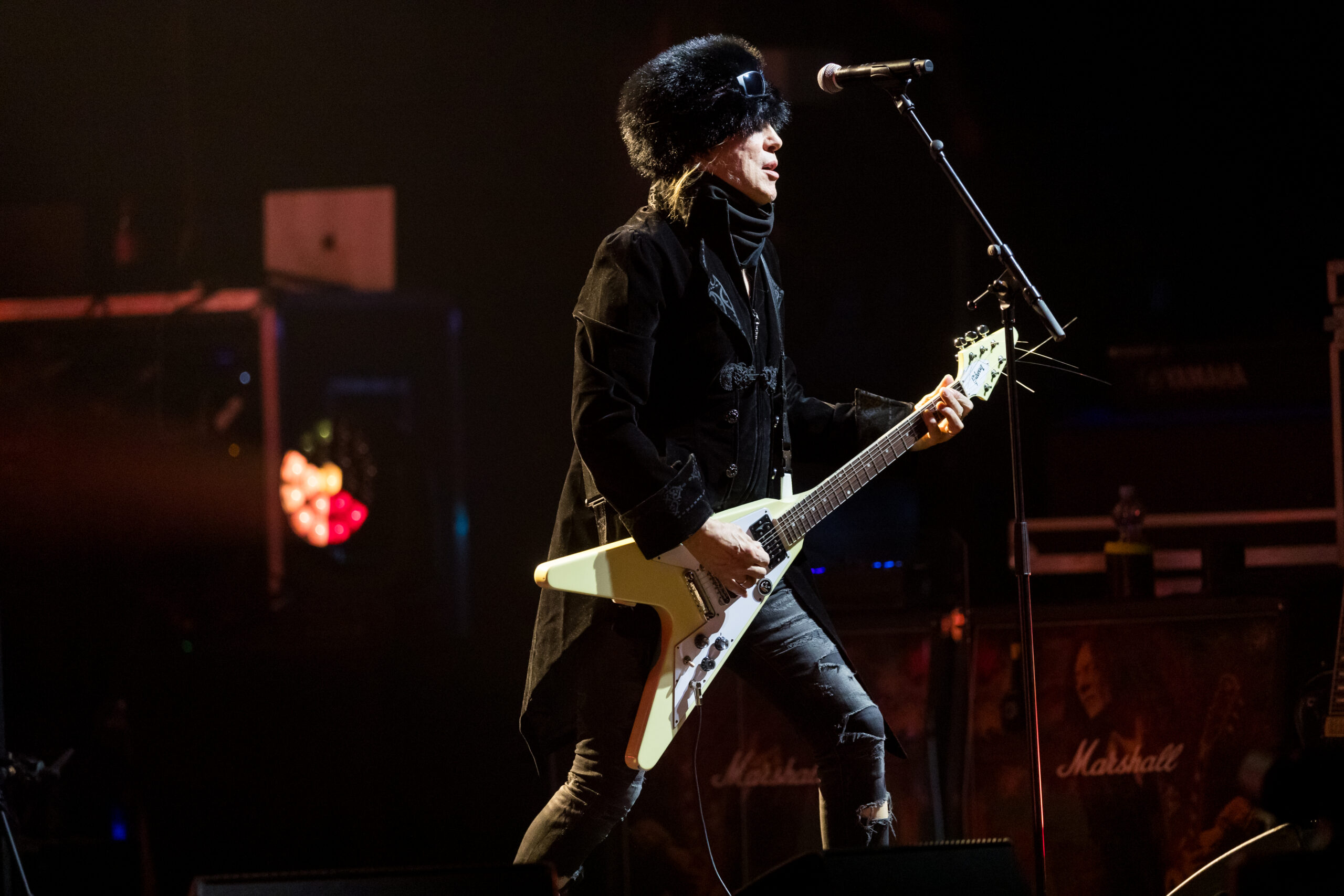 Michael Schenker playing a cream-colored Gibson Flying V onstage, with the full headstock and both V-shaped body tips visible.