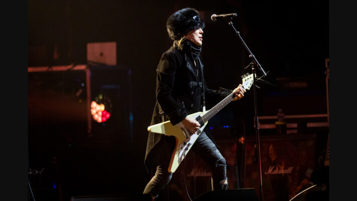 Michael Schenker plays a cream V-shaped electric guitar on a dark stage.