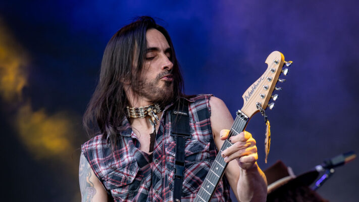 Nuno Bettencourt performing with Extreme, looking toward the guitar neck, with his full head and fretting hand visible against blue stage lighting.
