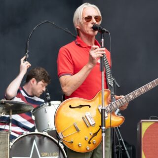 Paul Weller in sunglasses and a red polo shirt stands at a microphone with a sunburst semi-hollow electric guitar, drums and amplifiers behind him.