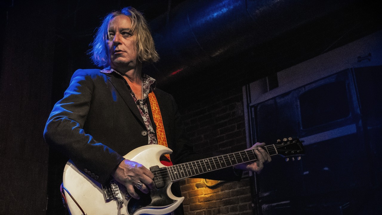Peter Buck plays a cream double-cut electric guitar on a dark blue-lit stage, with his full head, both hands and the guitar neck visible.