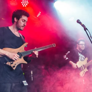 Plini playing a natural-finish headless electric guitar, with Simon Grove in the background, in a 2017 concert photograph.