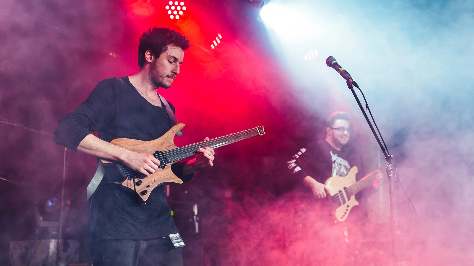 Plini playing a natural-finish headless electric guitar, with Simon Grove in the background, in a 2017 concert photograph.