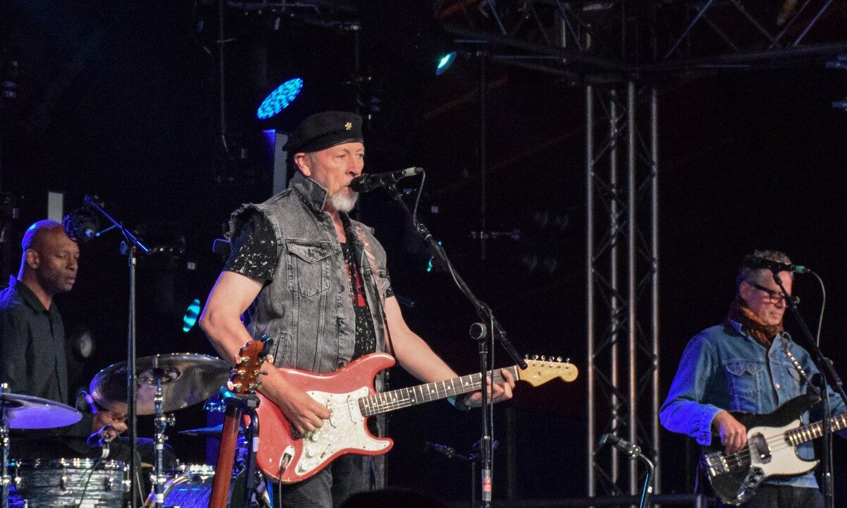 Richard Thompson in a black beret plays a red Stratocaster-style electric guitar onstage with his drummer and bassist.