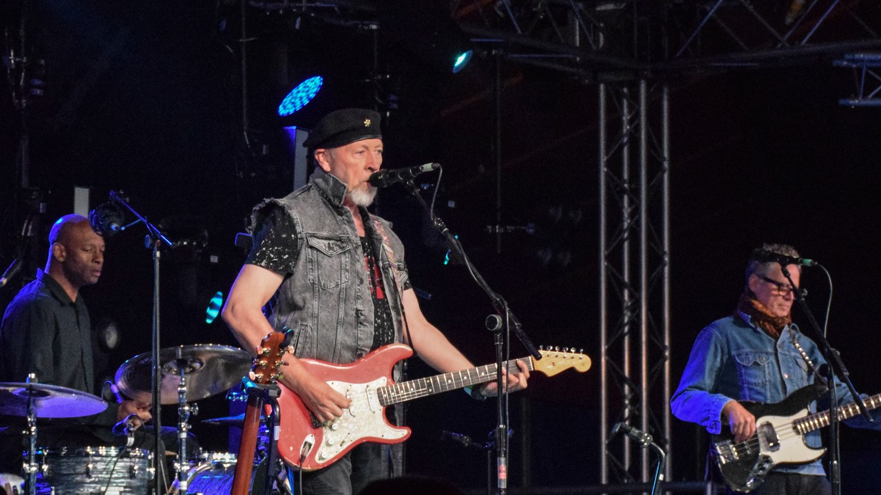 Richard Thompson in a black beret plays a red Stratocaster-style electric guitar onstage with his drummer and bassist.