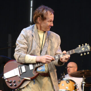 Robby Krieger playing a brown double-cut electric guitar, with his full head, both hands and complete guitar visible; dark margins preserve the whole photograph.