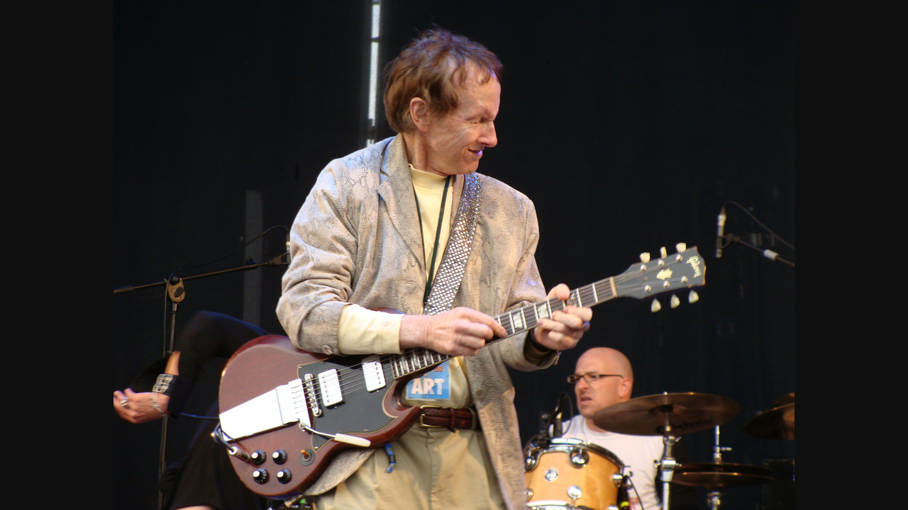 Robby Krieger playing a brown double-cut electric guitar, with his full head, both hands and complete guitar visible; dark margins preserve the whole photograph.