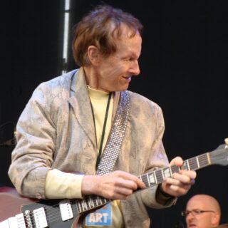 Robby Krieger playing a brown double-cut electric guitar, with his full head, both hands, neck and pickups visible.