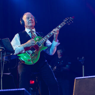 Robert Fripp seated on stage playing an electric guitar, with headphones on.