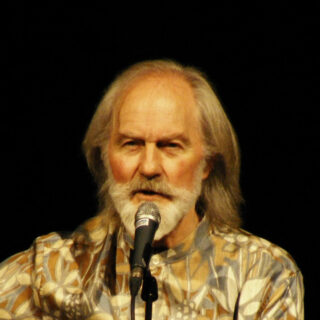 Roy Harper singing into a microphone in a patterned shirt, with his full head and beard visible against a dark stage background.