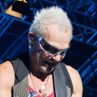 Rudolf Schenker performing with Scorpions in 2014, his full silver-haired head, sunglasses and upper torso visible against blue stage lighting.