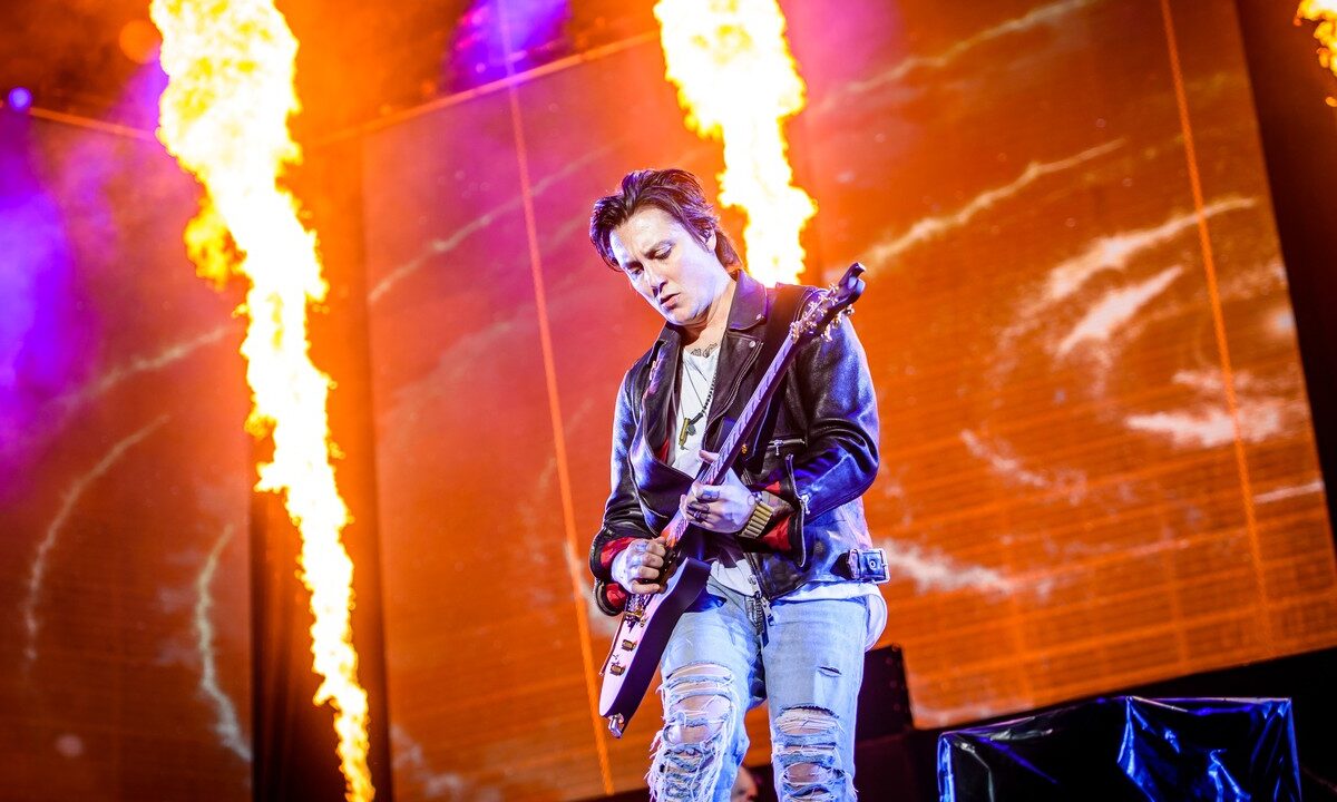Synyster Gates plays an angular electric guitar in front of orange stage flames, with his full head, both hands and the guitar visible.