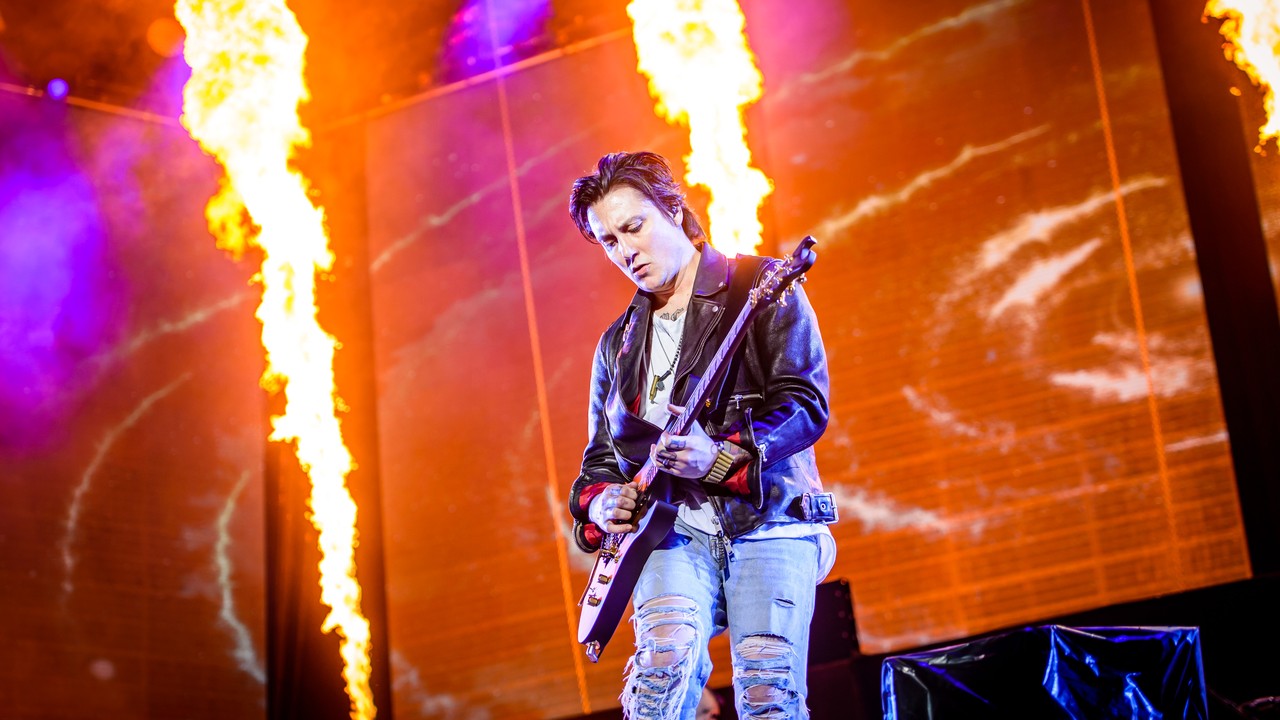 Synyster Gates plays an angular electric guitar in front of orange stage flames, with his full head, both hands and the guitar visible.