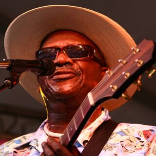 Taj Mahal singing into a microphone on stage in a floral shirt and straw hat, the headstock of his guitar in front of him, at the 2008 Houston International Festival.
