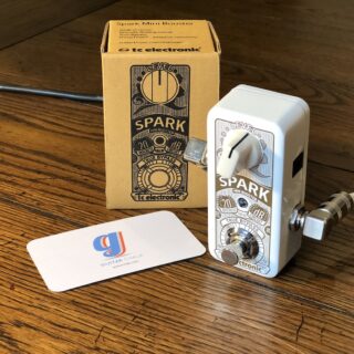 White TC Electronic Spark Mini Booster pedal with its original box on a wooden floor; the complete pedal, level knob and footswitch are visible.