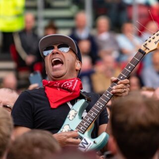 Tom Morello wearing sunglasses and a red bandana while playing a pale blue electric guitar among a concert crowd.