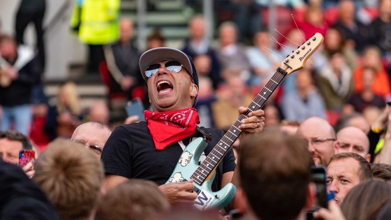 Tom Morello wearing sunglasses and a red bandana while playing a pale blue electric guitar among a concert crowd.