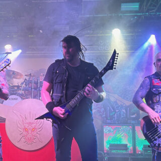 Trivium performing onstage under colored lighting, with three musicians and their instruments visible.