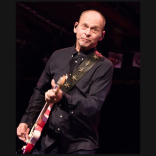 Wayne Kramer playing guitar live, with head and both hands visible