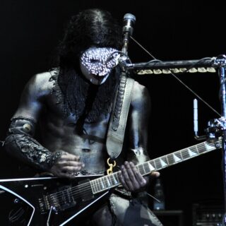 Wes Borland in his stage costume, playing an angular black electric guitar; his head, hands and most of the guitar are visible.